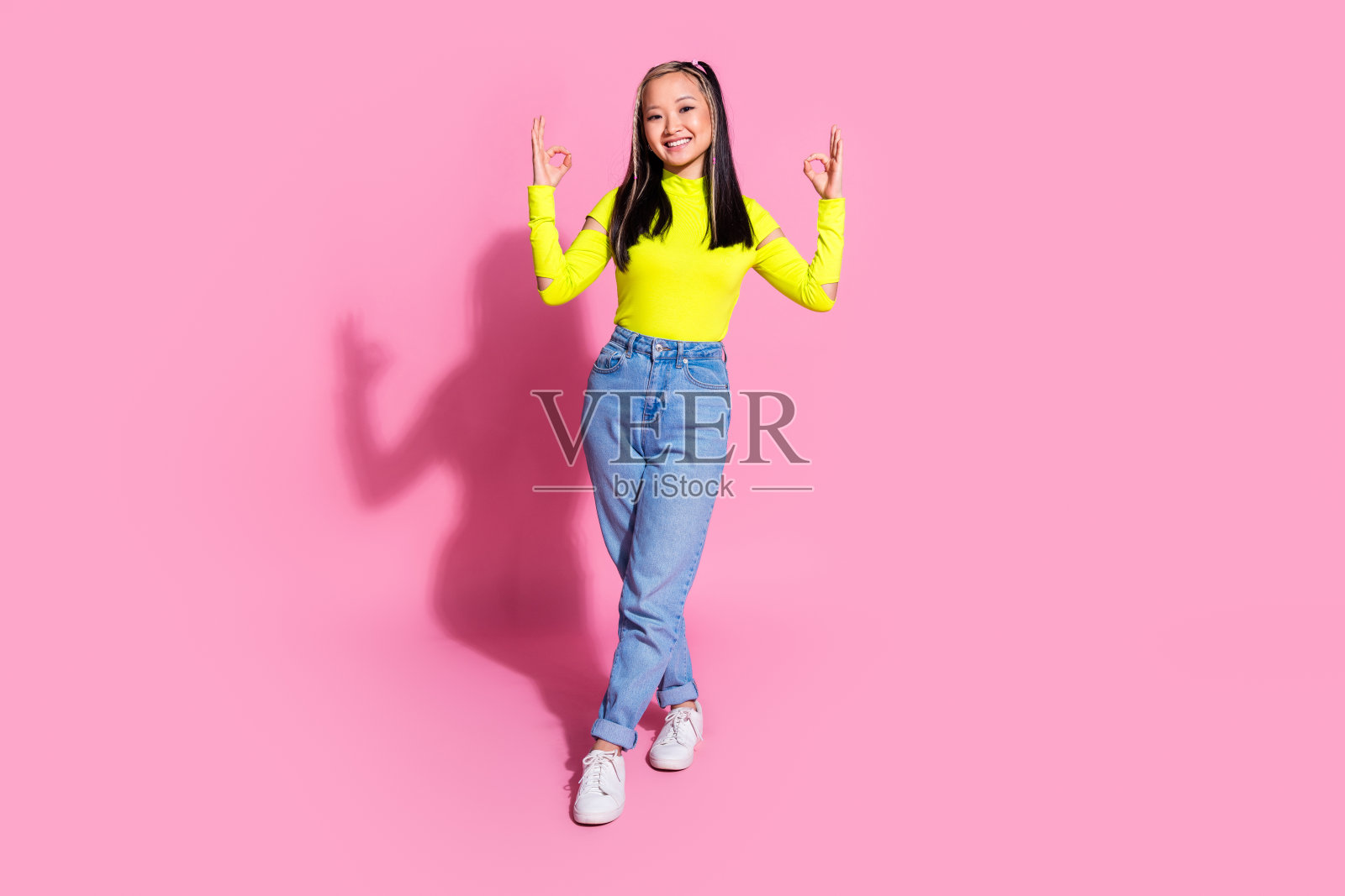 满意乐观的漂亮女孩的全身照片,穿着黄色衬衫牛仔裤,在粉红色的背景上显示出okey符号照片摄影图片
