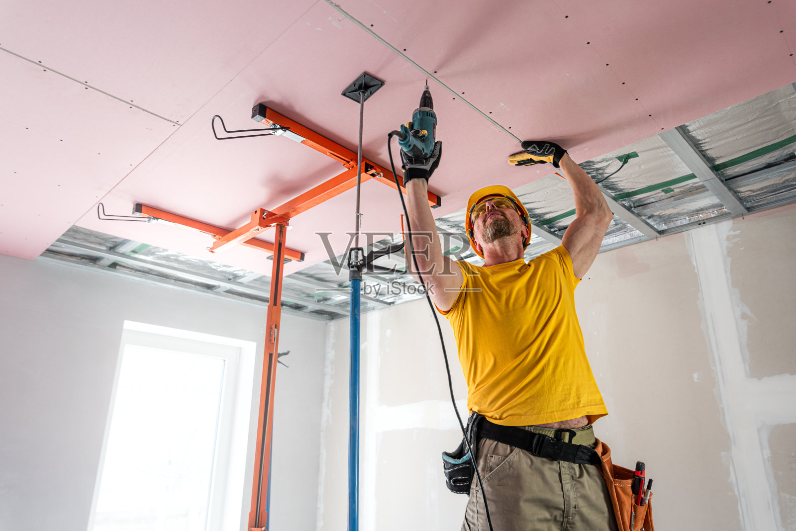 工人把石膏板拧到天花板上。他正在使用特殊的电动螺丝刀。红色石膏板提高天花板结构的防火性能。照片摄影图片