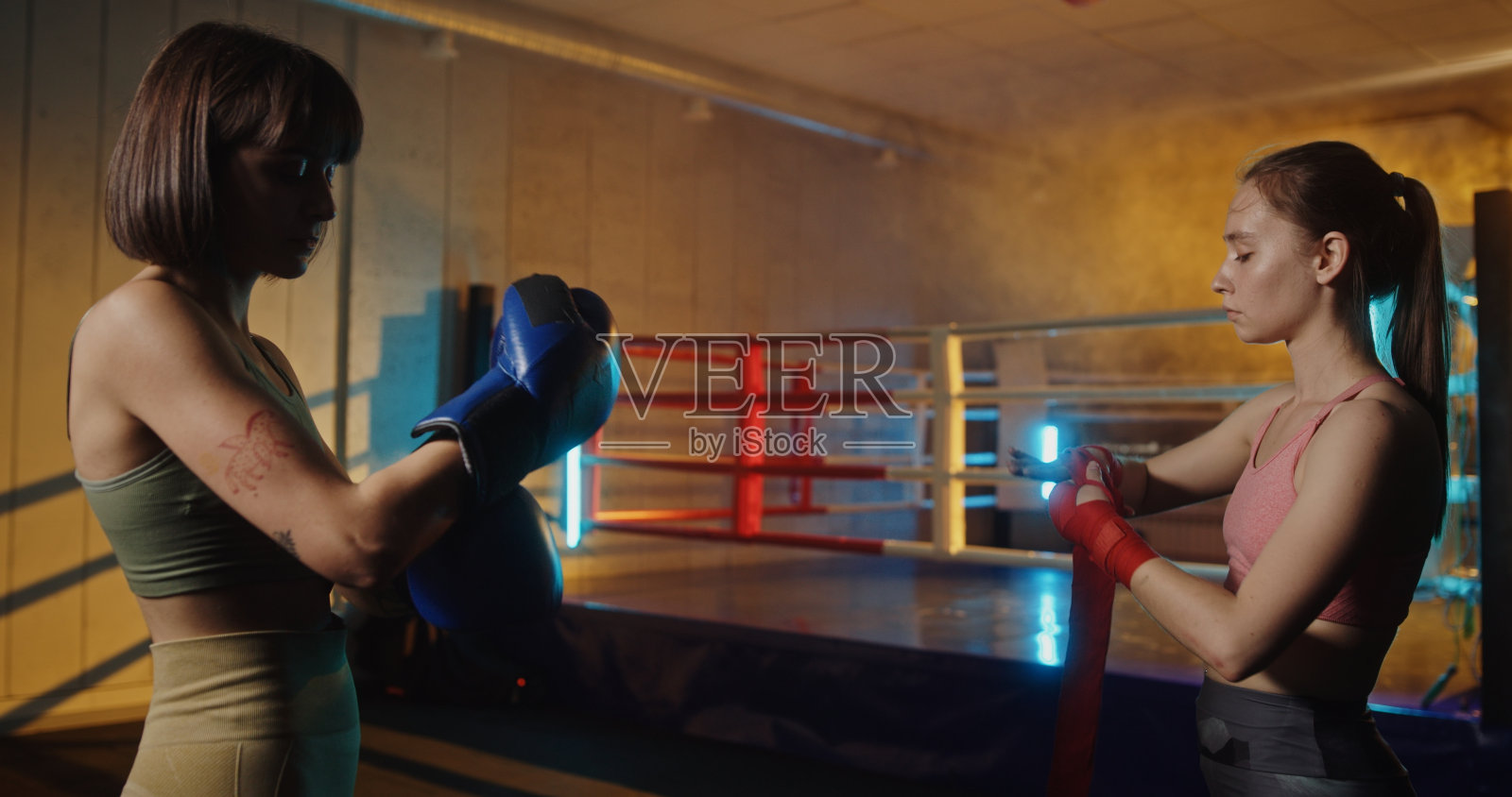 两名年轻女子站在拳击台前,双手缠上绷带,准备参加拳击比赛。她们在彩色灯光的现代健身房里强调健身、训练和积极的生活方式照片摄影图片
