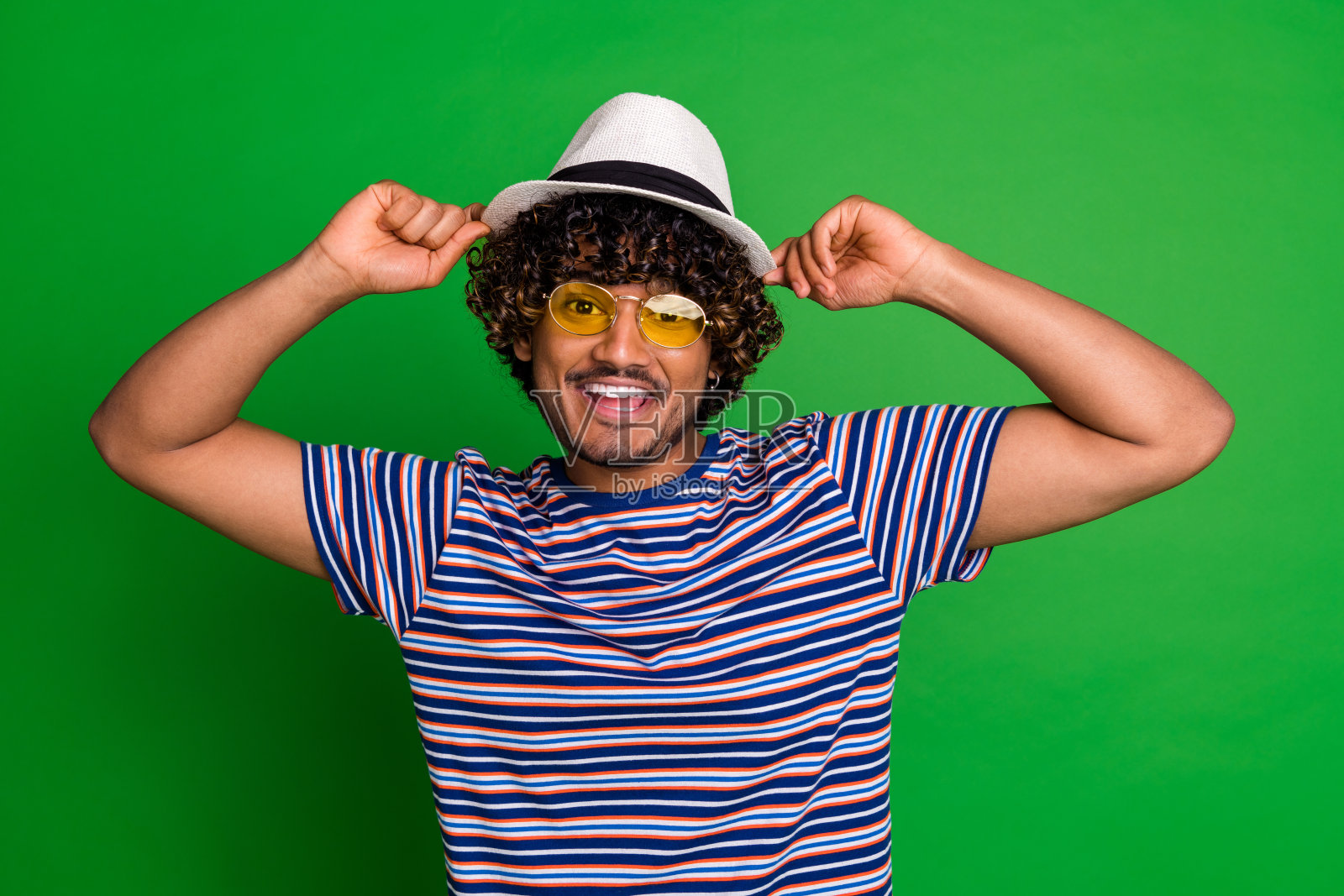 年轻的墨西哥酷酷的卷发滑稽的男人戴着太阳镜和遮阳帽学生条纹t恤孤立的绿色背景的肖像照片摄影图片