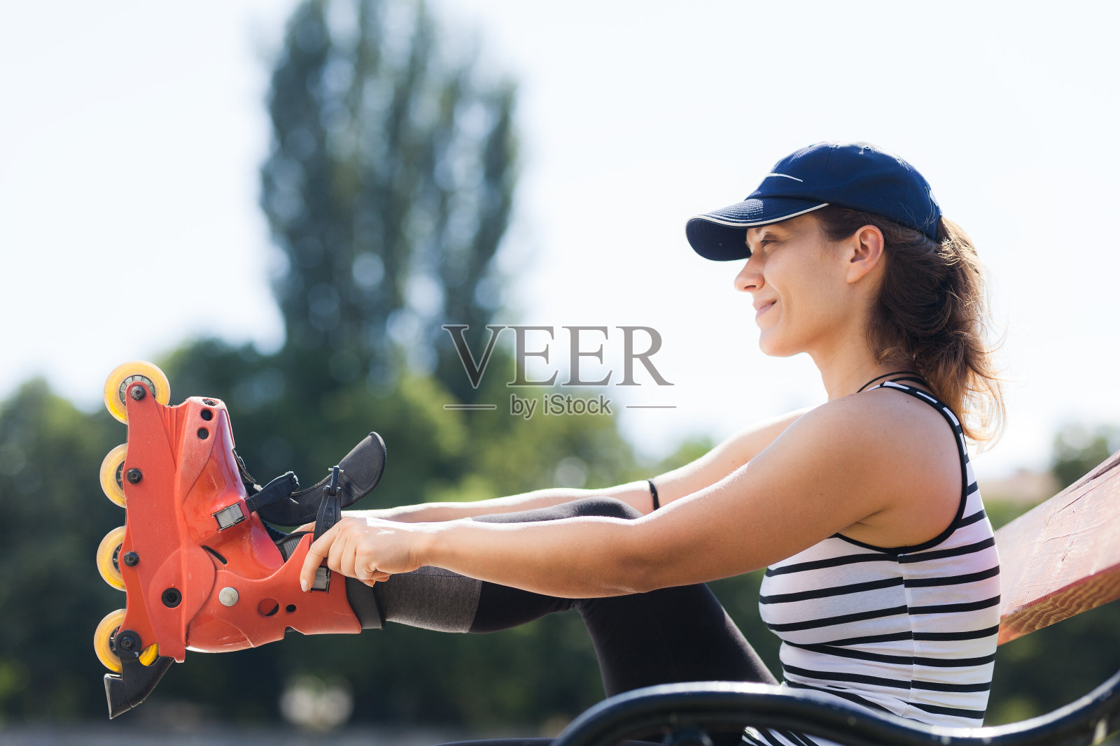 在一个阳光明媚的公园里,一位穿着条纹背心和鸭舌帽的微笑女子坐在长凳上,在滑旱冰之前试穿她的单排冰鞋。照片摄影图片