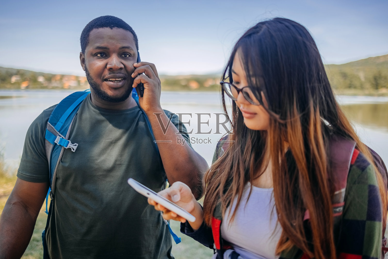年轻的多种族夫妇一起在湖边徒步旅行,同时使用手机照片摄影图片