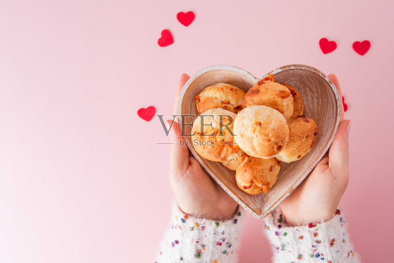 一个女孩的手拿着一个巴西奶酪面包,在柔和的粉红色背景上,有红色的小心脏和一个心形的碗照片摄影图片