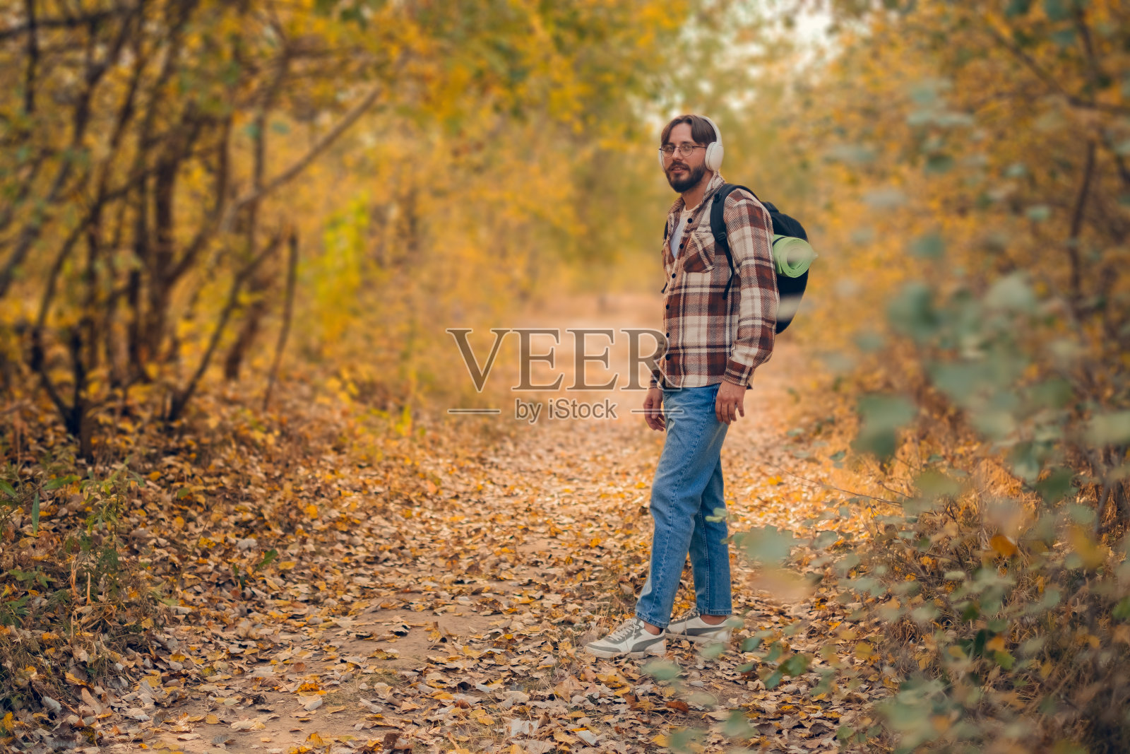 一个男人在森林里徒步,背着背包,戴着无线耳机,一边听着有声读物,一边沿着小路走。照片摄影图片