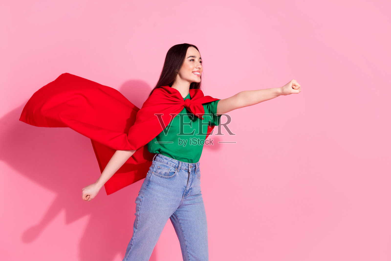 照片中开朗漂亮的女士穿着绿色的t恤,红色的斗篷升起,拳头空着,孤立的粉红色背景照片摄影图片