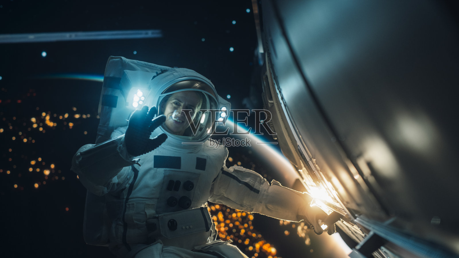 在宇宙飞船、卫星或空间站外太空行走时,一位美丽的宇航员穿着宇航服摆姿势的肖像。女宇航员看着镜头,微笑着用手挥手打招呼照片摄影图片