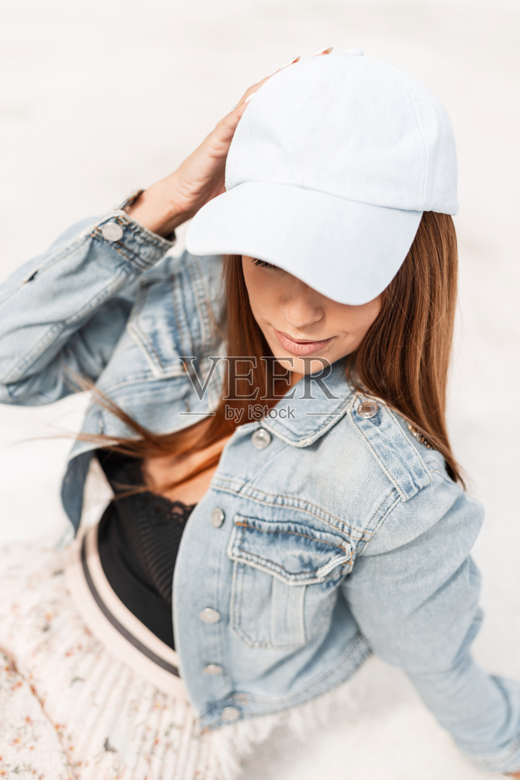 时尚漂亮的年轻女子穿着时尚的牛仔夹克,戴着蓝色的帽子,坐在白色的沙滩上休息照片摄影图片