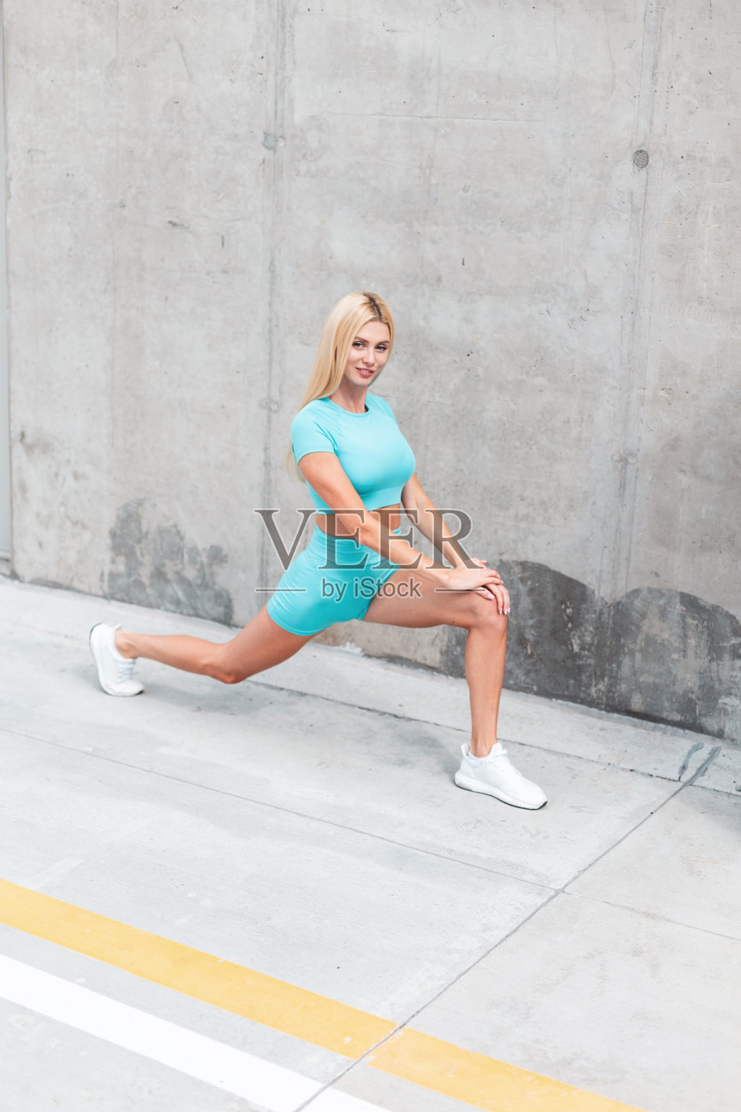 年轻漂亮的健身模特穿着蓝色运动服和白色运动鞋在街上的一堵灰色的墙旁边锻炼和伸展照片摄影图片