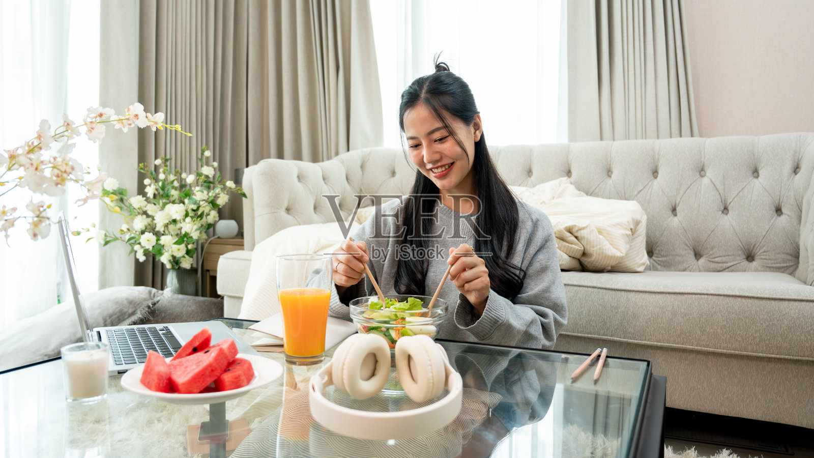 一个积极、迷人的年轻亚洲女人在客厅的咖啡桌旁吃着健康的沙拉。照片摄影图片