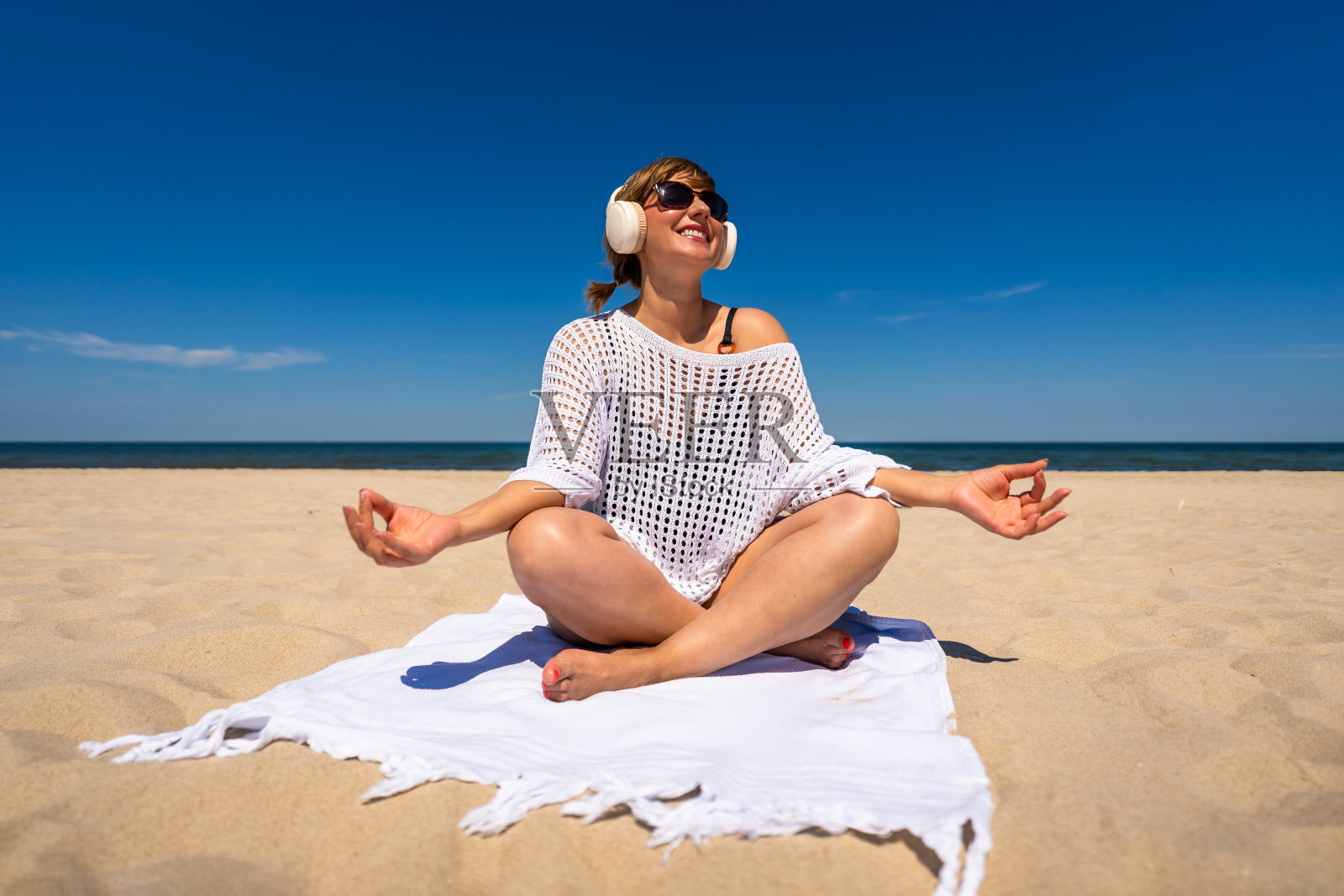 暑假在沙滩上听音乐。美丽的年轻女子穿着黑色泳衣和白色束腰外衣,戴着耳机听着音乐,盘腿坐在白色沙滩上的毛巾上。前视图照片摄影图片