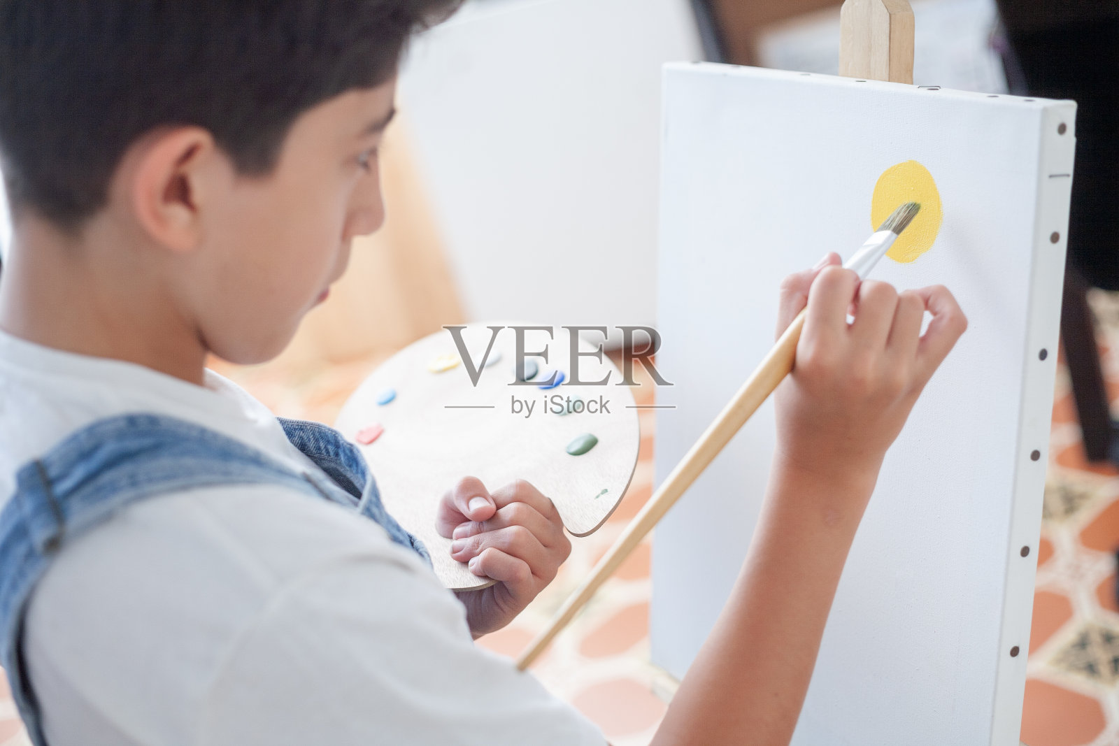 一个男孩正在白色画布上画画。他使用画笔和调色板上的各种颜色。照片摄影图片