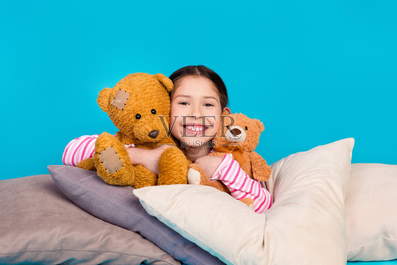 照片中,友善开朗的小女孩穿着条纹衬衫,躺在枕头上,抱着毛茸茸的泰迪熊,背景是蓝色的照片摄影图片