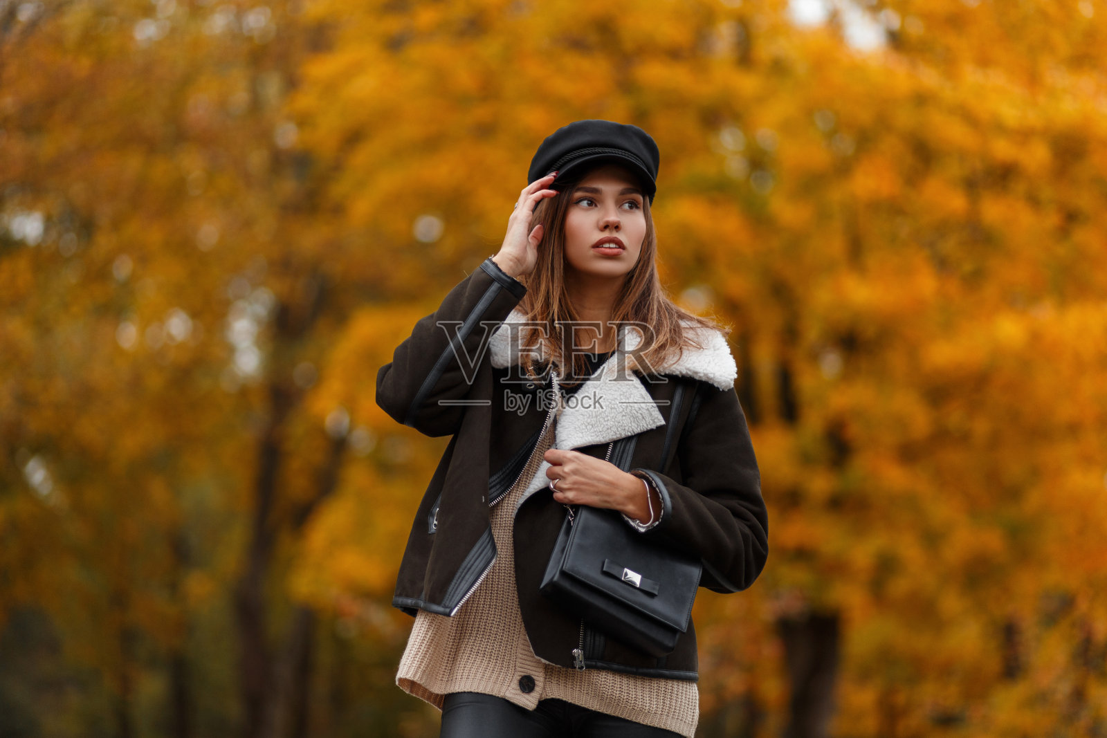 欧洲年轻女子穿着时髦的衣服,戴着复古的帽子,提着黑色的皮包,在公园里摆姿势。迷人的时尚女孩时装模特在森林的背景金树叶。秋天的风格。照片摄影图片
