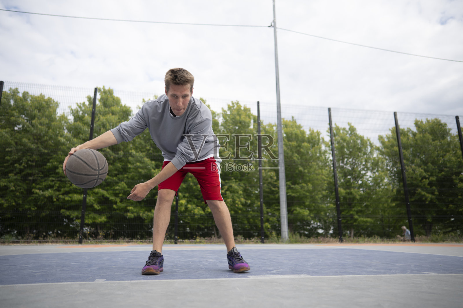 一个年轻人正在室外球场练习篮球运球。他穿着灰色的运动衫和红色的短裤照片摄影图片