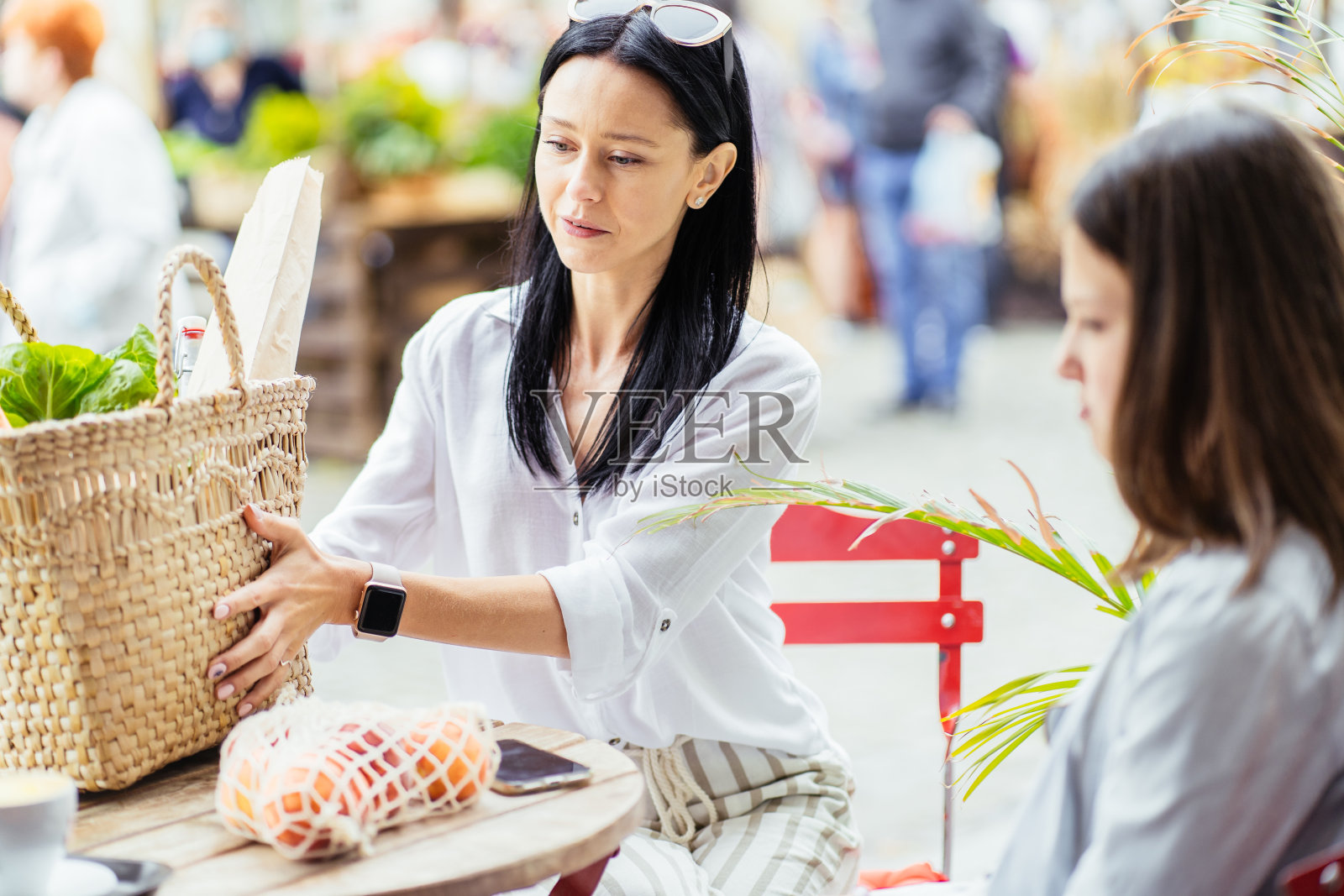 黑发苗条的母亲和她十几岁的女儿在农贸市场购物后放松,坐在户外咖啡馆里,柳条袋里装满了水果,绿色,蔬菜。照片摄影图片
