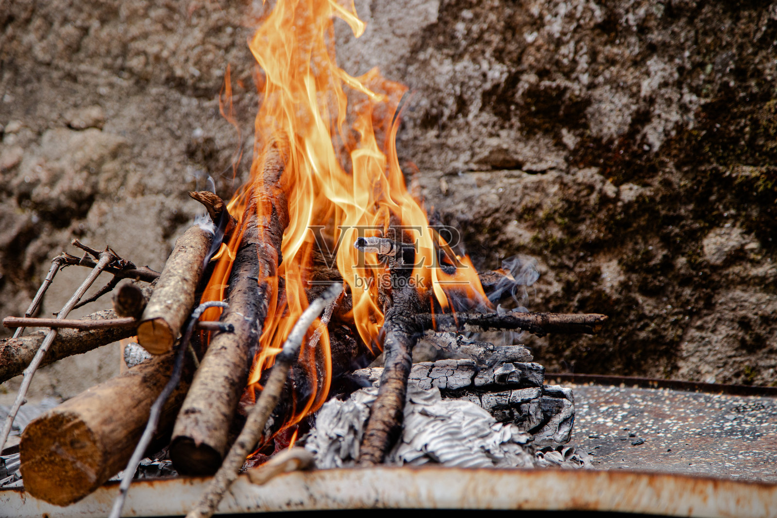 一个小小的户外火坑,靠在质朴的石墙上,用原木和引火点燃,发出明亮的光芒,非常适合在露营旅行或户外聚会期间做饭或取暖。照片摄影图片