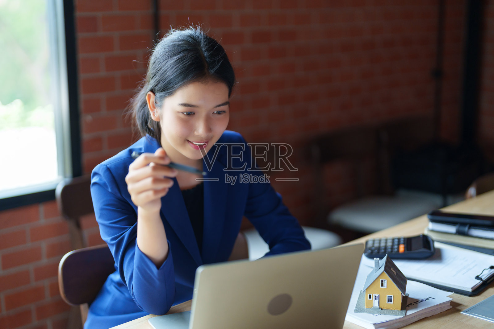 一位身穿蓝色西装的卖房人正在办公桌前用笔记本电脑工作。背景是一面砖墙。在桌子上,有一个小样板房和一些书。照片摄影图片