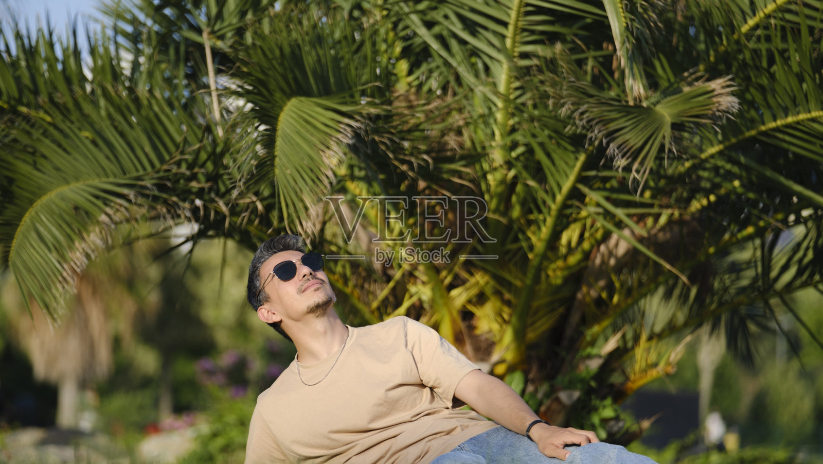 一个英俊、年轻、黑发的男人懒洋洋地躺在一个树木繁茂的公园的草地上,摆着很酷的姿势,享受着自己照片摄影图片