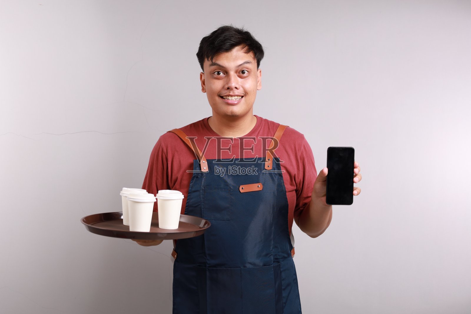 年轻快乐的亚洲人咖啡师调酒师员工穿着蓝围裙在咖啡店工作拿着纸杯手机空白屏幕区域孤立在白色背景上,创业照片摄影图片