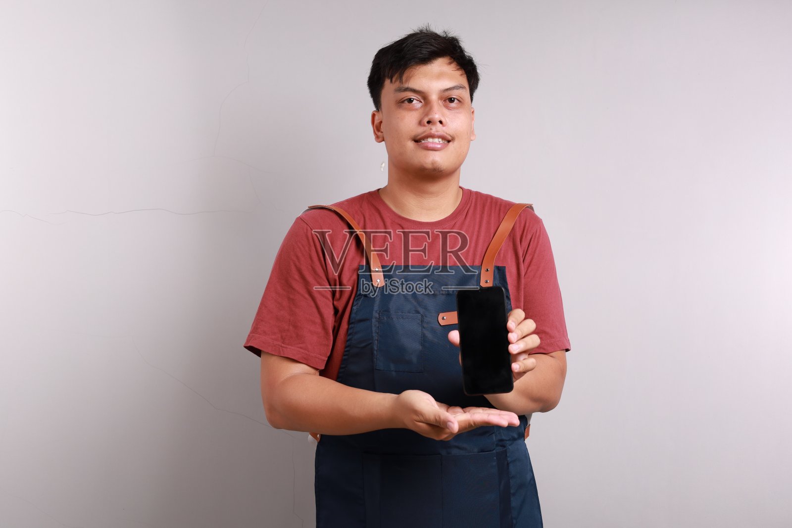 穿着蓝色围裙、微笑着的亚洲酒保在白色背景上展示着空白的智能手机屏幕照片摄影图片