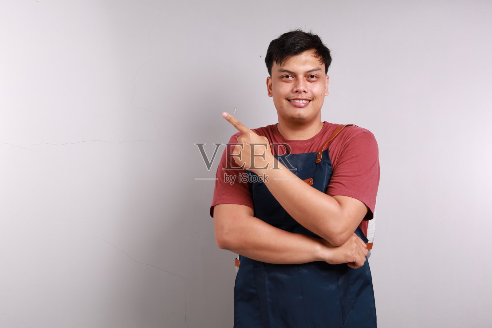 快乐的亚洲咖啡师指着左边微笑着,穿着蓝色围裙制服,站在白色的背景下。本空间照片摄影图片