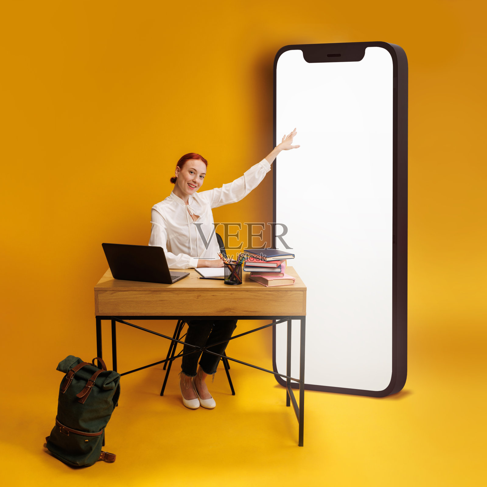 教师工作区。年轻人指着巨大的智能手机屏幕,坐在桌子前,学习用品和笔记本电脑在黄色的背景下照片摄影图片