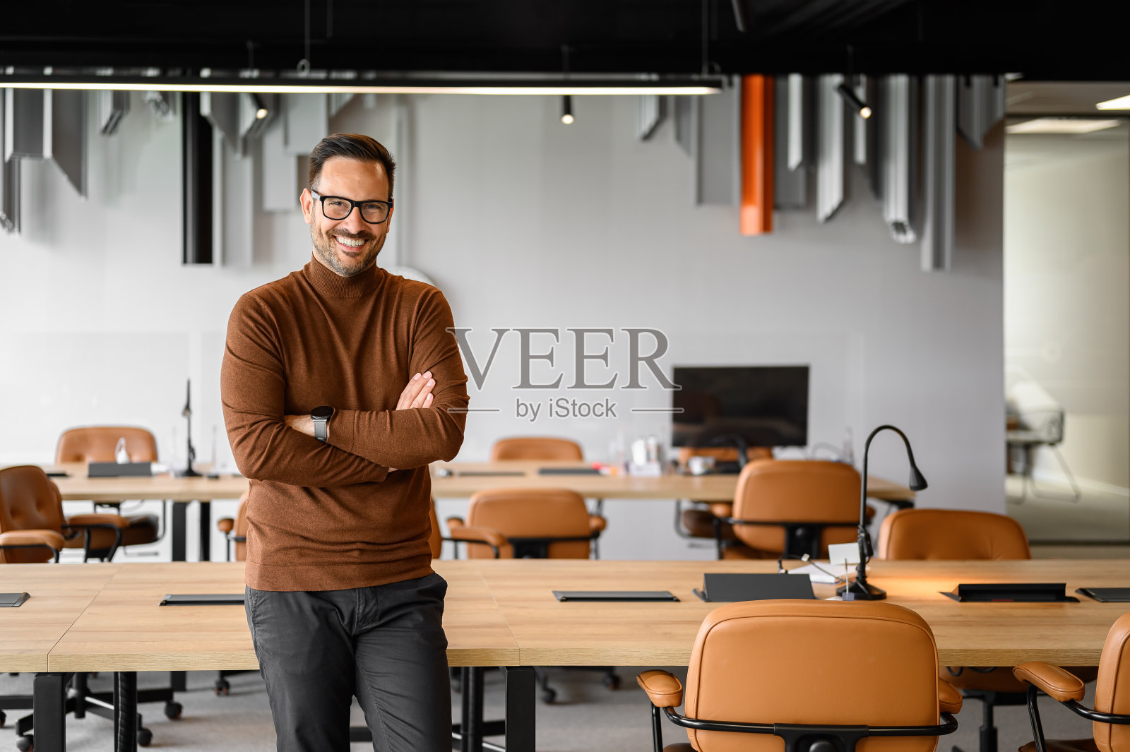 英俊的年轻人双臂交叉,微笑着站在办公室的会议桌和椅子旁照片摄影图片