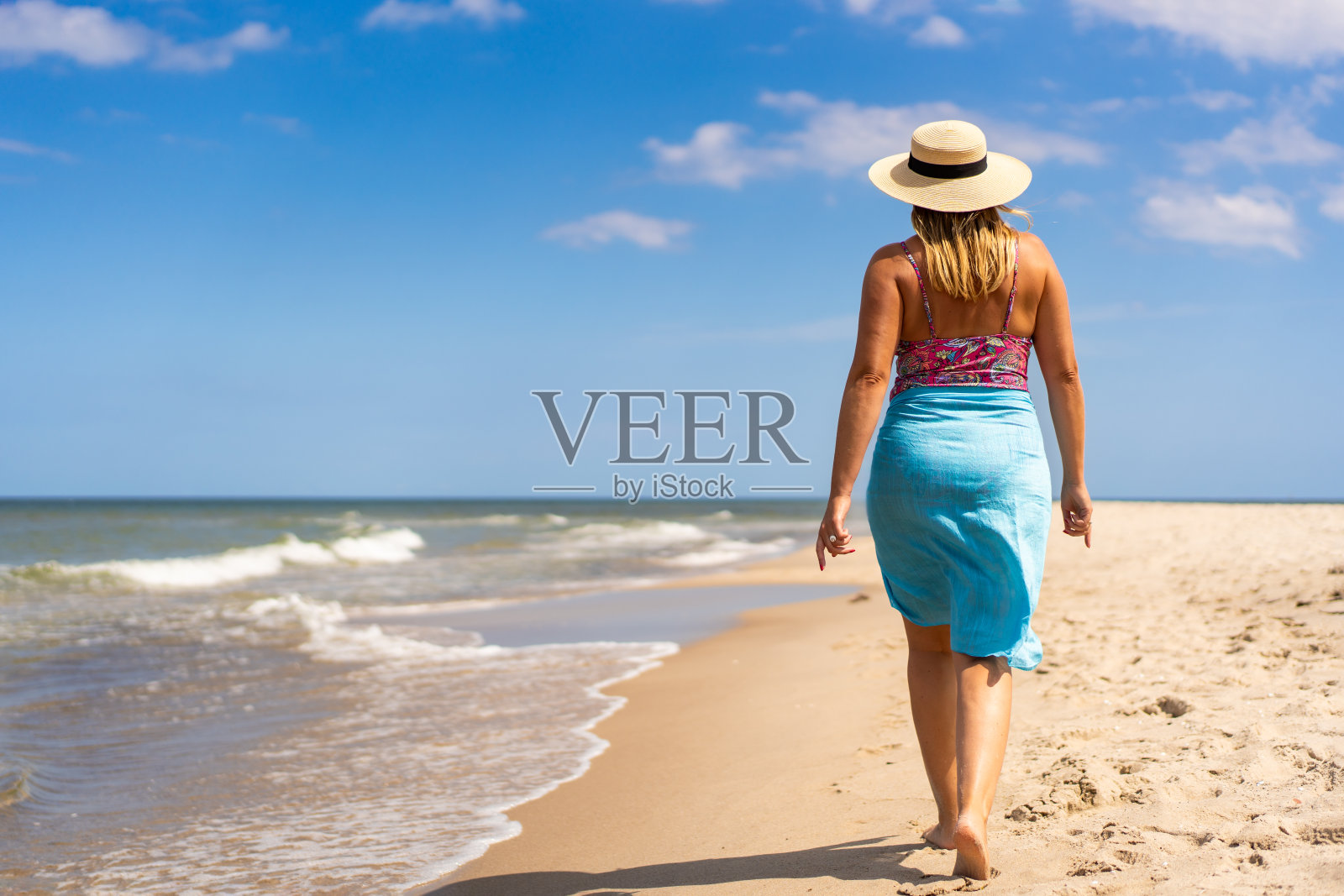 暑假。美丽的年轻女子在一个美丽的阳光明媚的日子里,穿着五颜六色的泳衣,戴着蓝色的帽子,走在沙滩上。后视图照片摄影图片