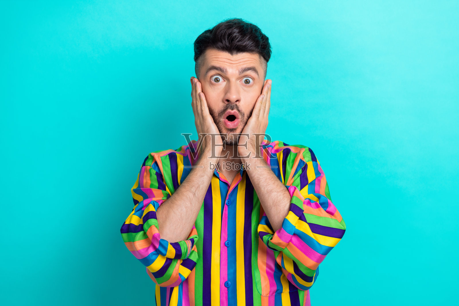 好心情的肖像令人印象深刻欣喜若狂的家伙,深色头发,穿着彩色衬衫,手掌放在颧骨上,孤立在蓝绿色的背景上照片摄影图片