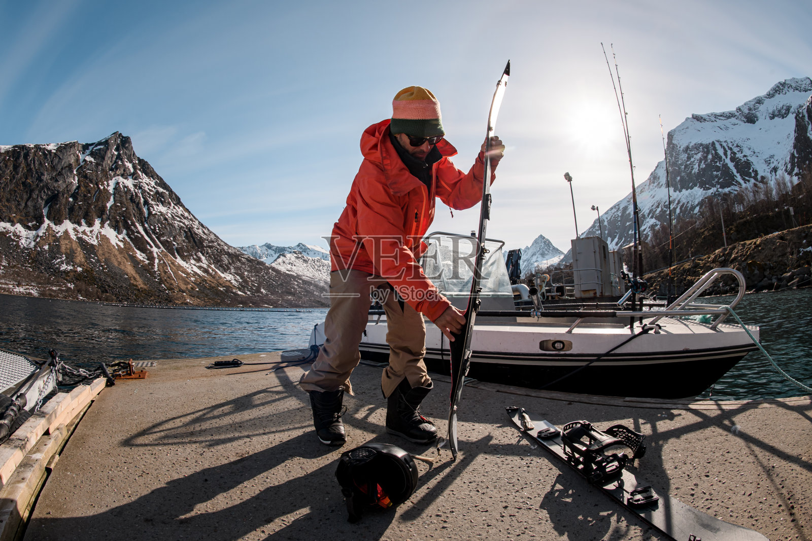 一名身穿橙色夹克的男子站在码头上的一艘渔船旁,调整着他的滑雪板照片摄影图片