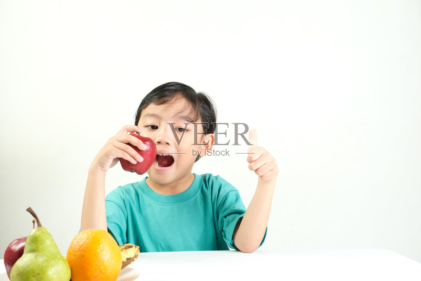 一个穿着青色衬衫的快乐男孩咬了一口红苹果,竖起大拇指。照片摄影图片