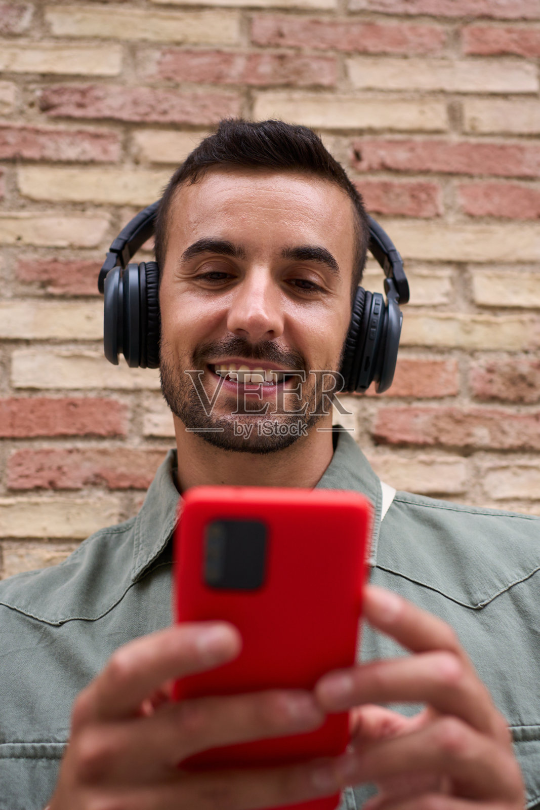 近距离拍摄靠在砖墙上使用红色手机的迷人年轻人。沉迷于技术。照片摄影图片