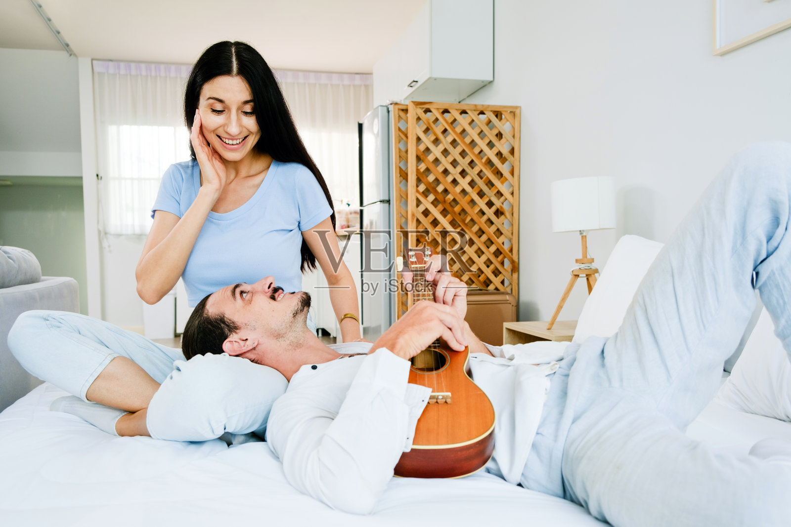 可爱的白人夫妇一起享受音乐。一个男人躺在床上一个女人的腿上弹奏着尤克里里琴,一个女人在卧室里欢快地唱歌。照片摄影图片