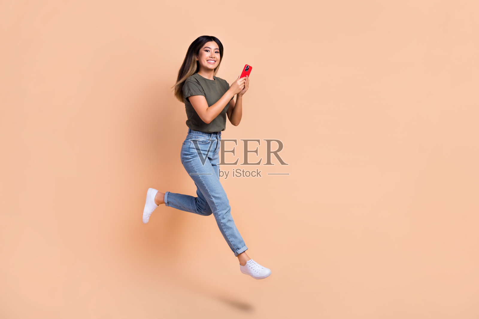 时髦跳跃匆忙的韩国女子用智能手机浏览互联网技术的全身照片,以米色为背景照片摄影图片