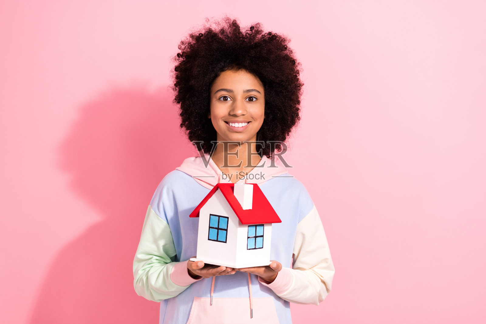 照片中开朗漂亮的小孩穿着连帽衫微笑着抱着小房子孤立的粉红色背景照片摄影图片