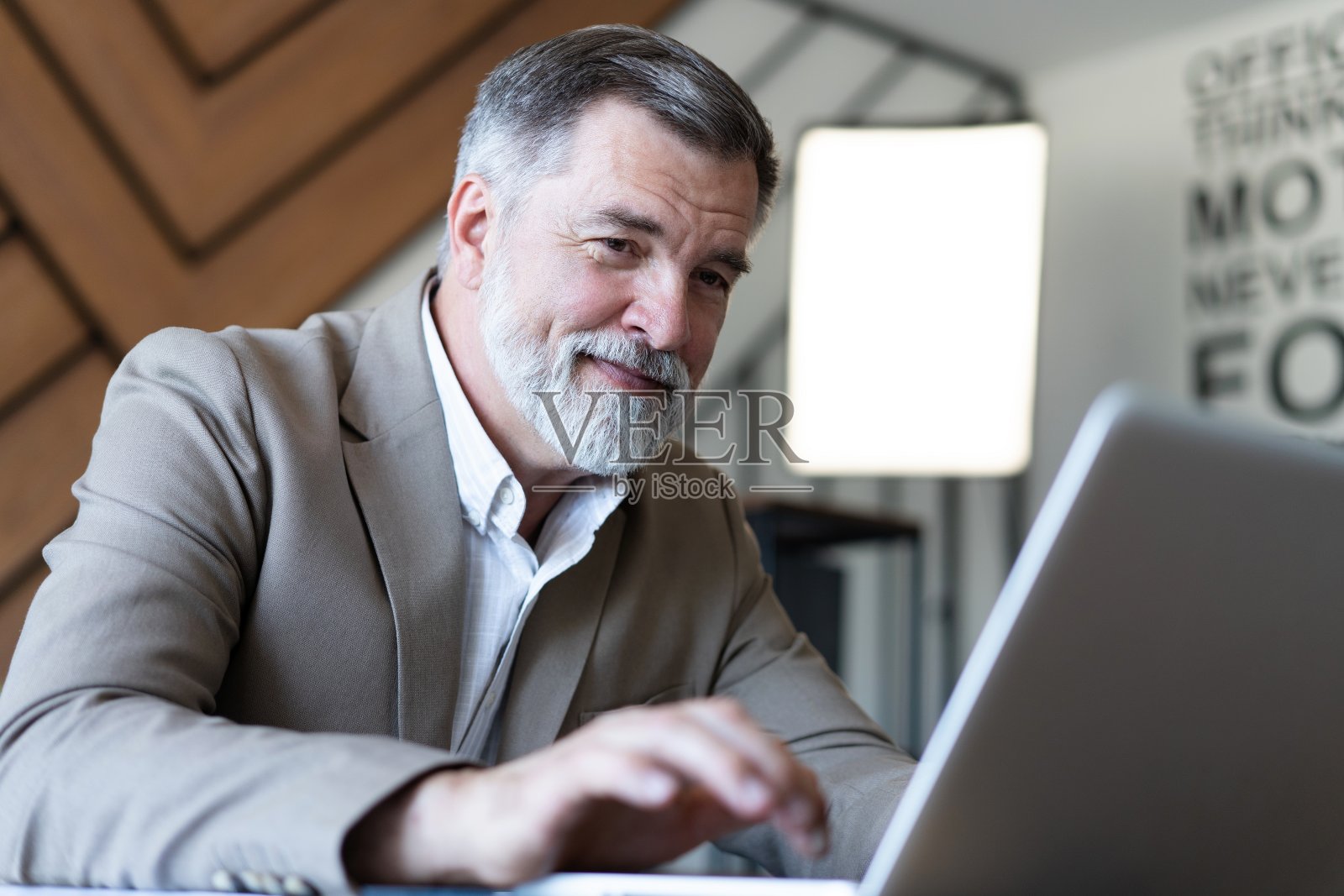 微笑的成熟的商人,穿着衬衫,坐在办公桌前使用笔记本电脑。快乐忙碌的职业中年商人投资者在电脑上工作,在办公室里望着远方照片摄影图片