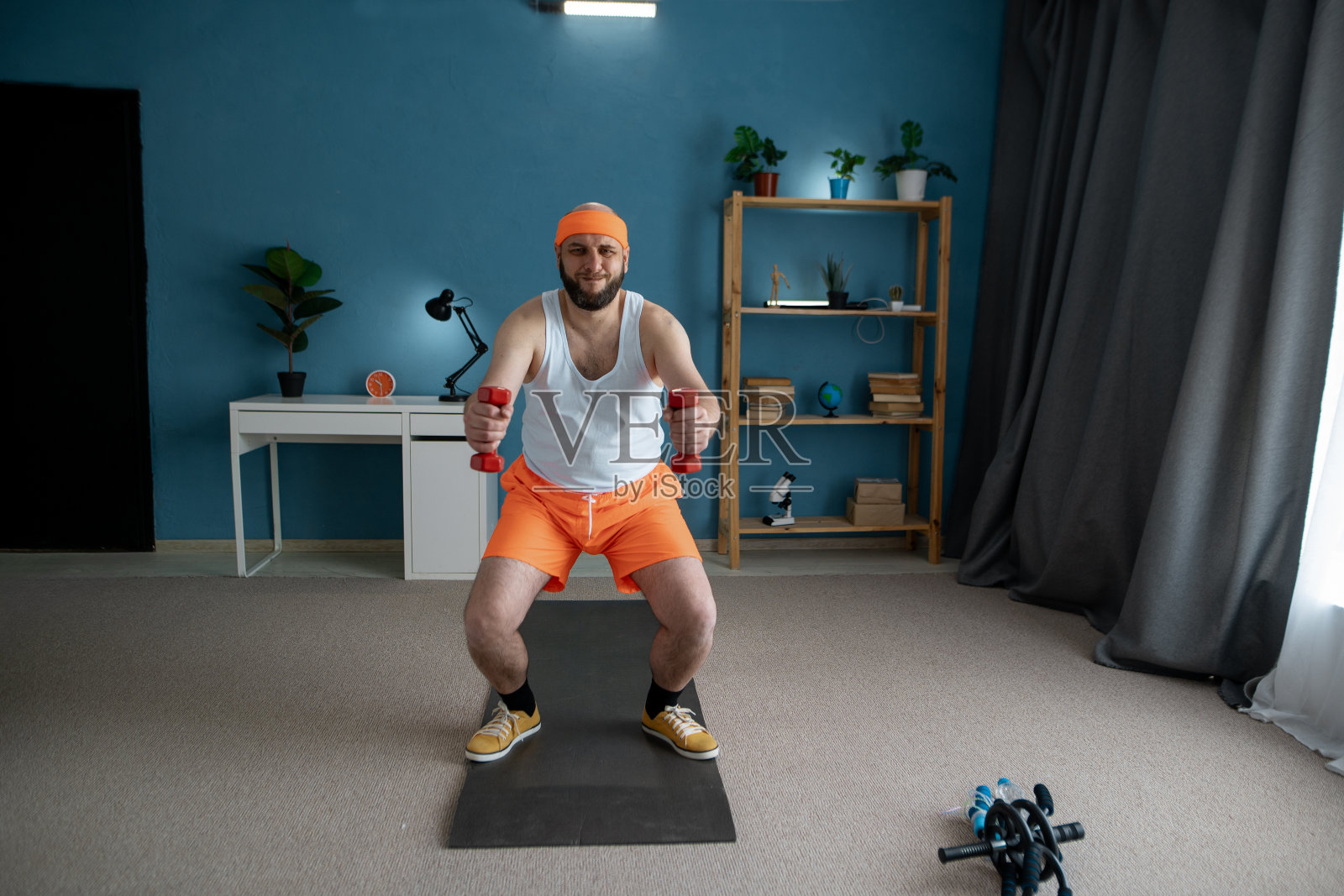 滑稽的人在家里用哑铃做运动。男运动员稍微坐下,双臂向前伸展,手持两个哑铃照片摄影图片