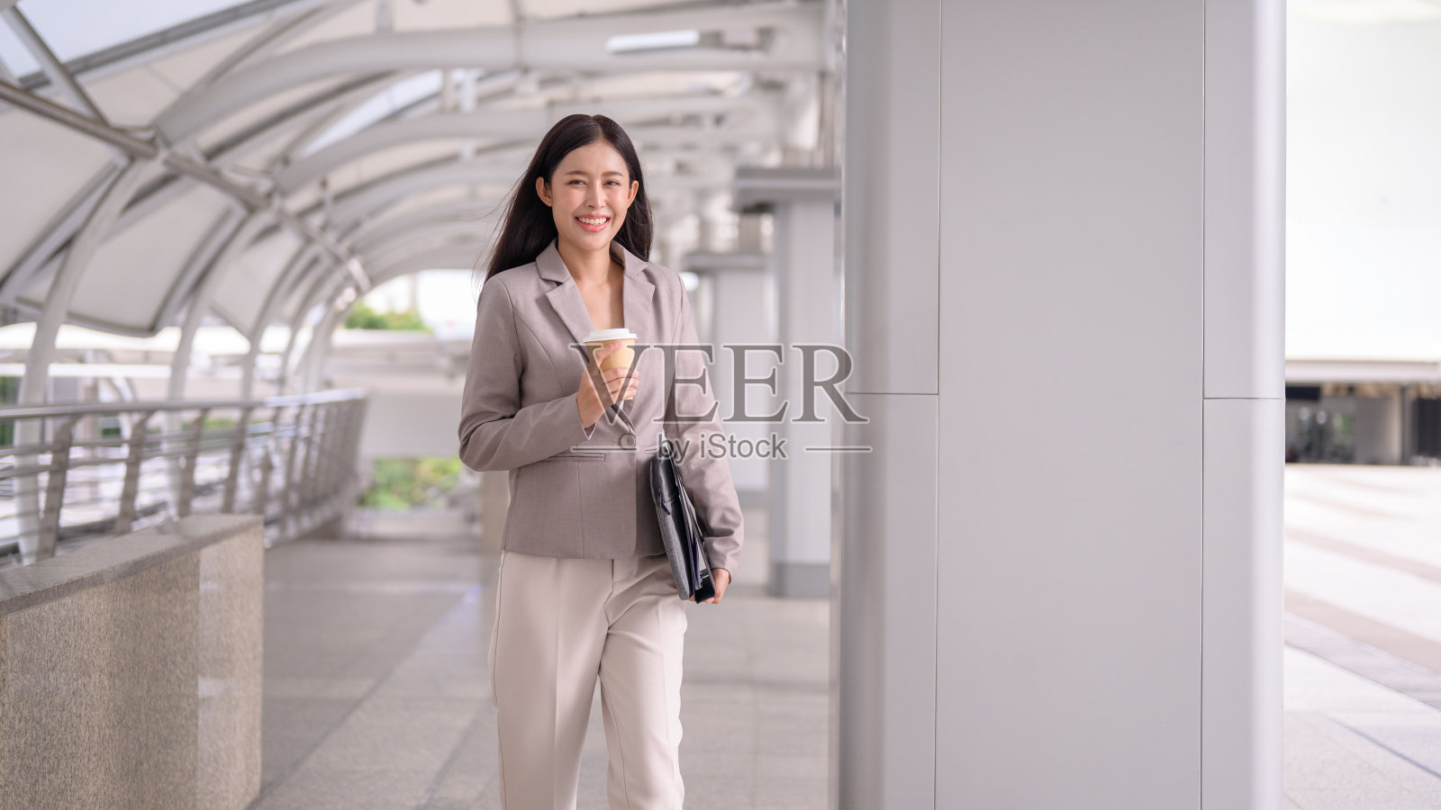 微笑的年轻女商人端着一杯咖啡和一个黑色文件夹,自信地走在现代都市环境中。照片摄影图片