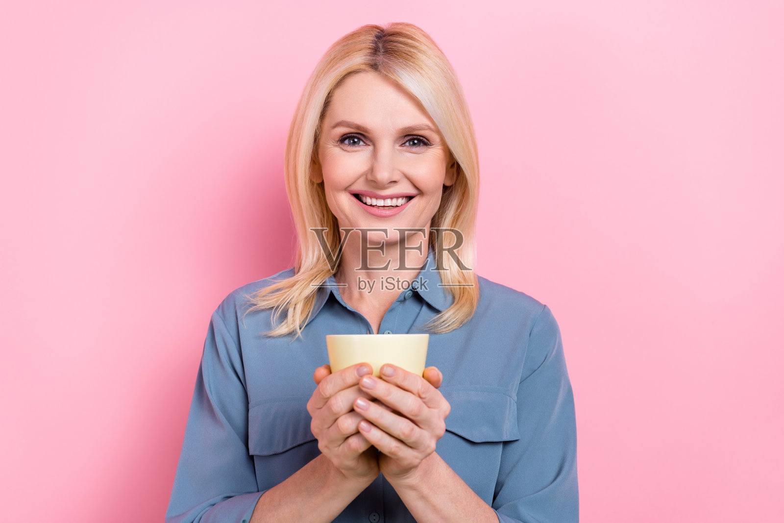 迷人的高级商务女士的照片拿着热杯咖啡休息时间穿着聪明休闲的蓝色衬衫孤立的粉红色背景照片摄影图片