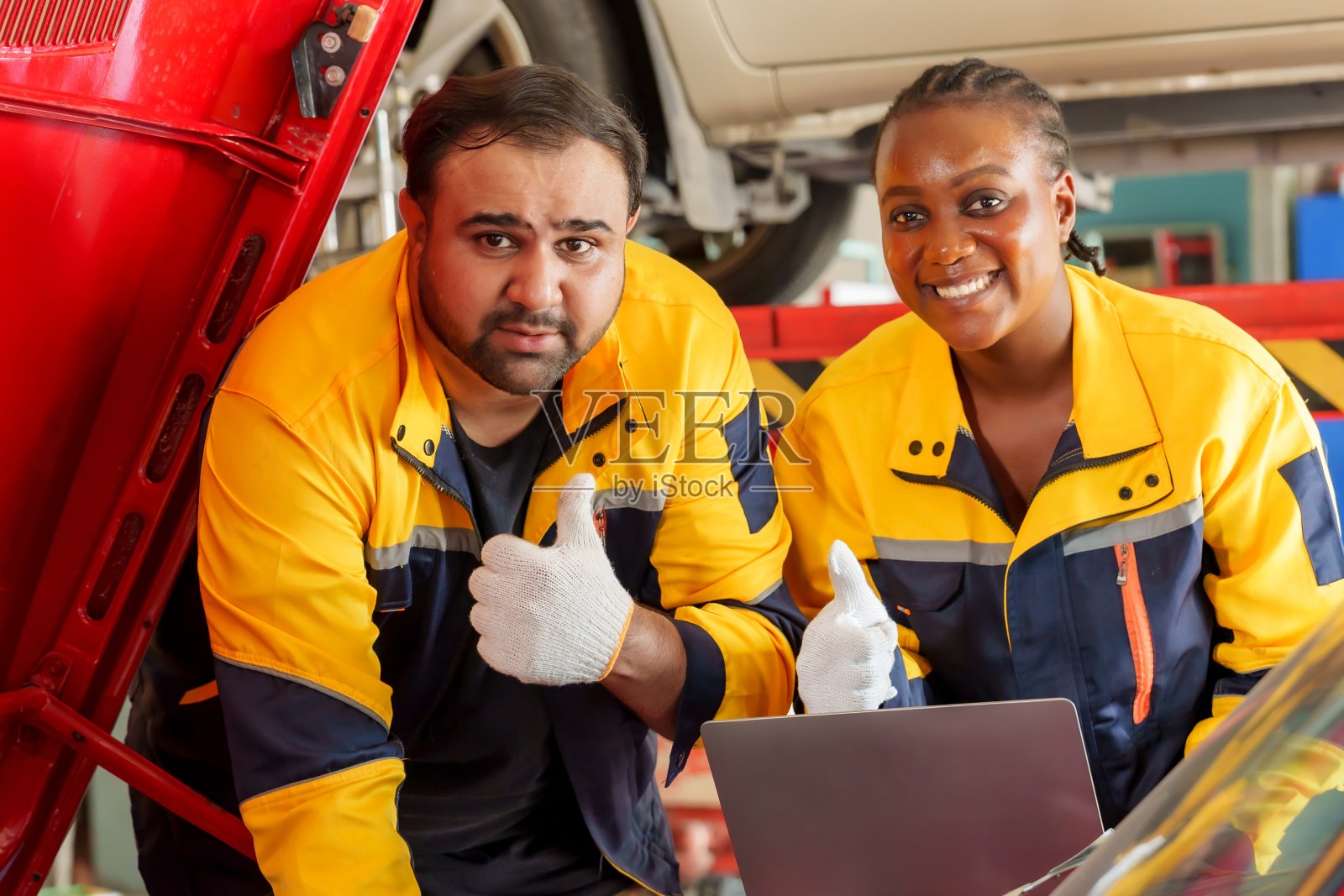 男技工和女技工微笑着竖起大拇指,在车库里修理一辆红色的汽车发动机。穿着黄色和蓝色的制服。强调满意度,团队合作和成功的修复结果照片摄影图片