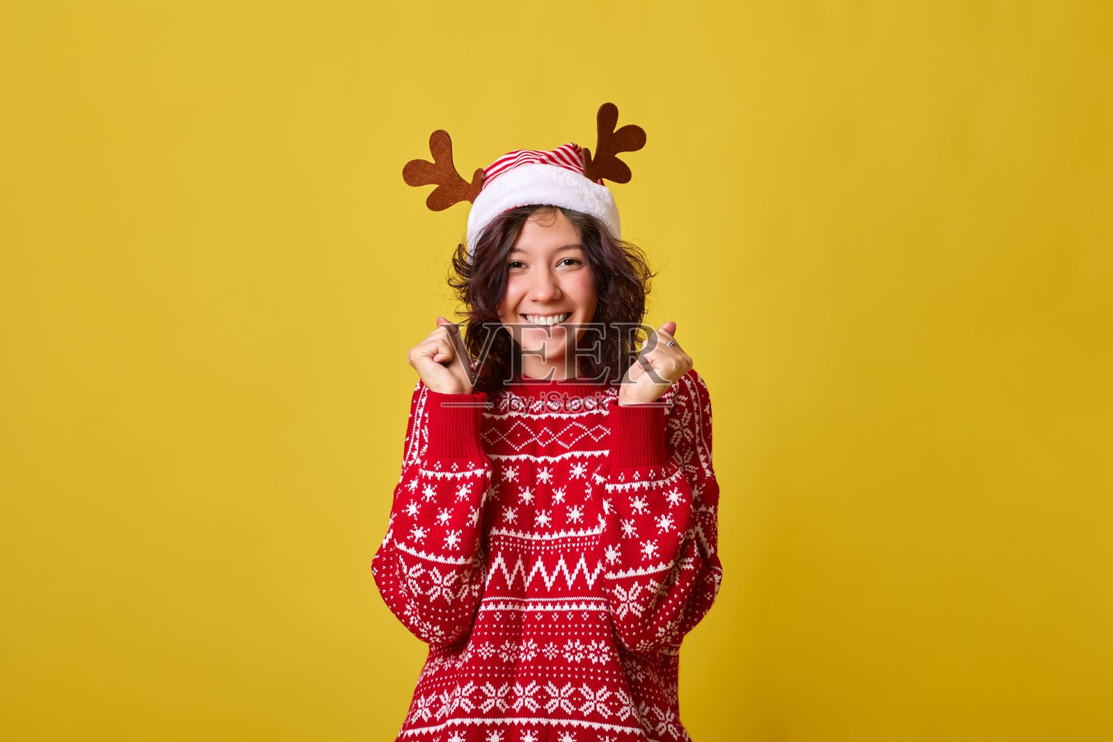 开朗的年轻女子戴着圣诞老人帽,黄色画室背景上有驯鹿角照片摄影图片