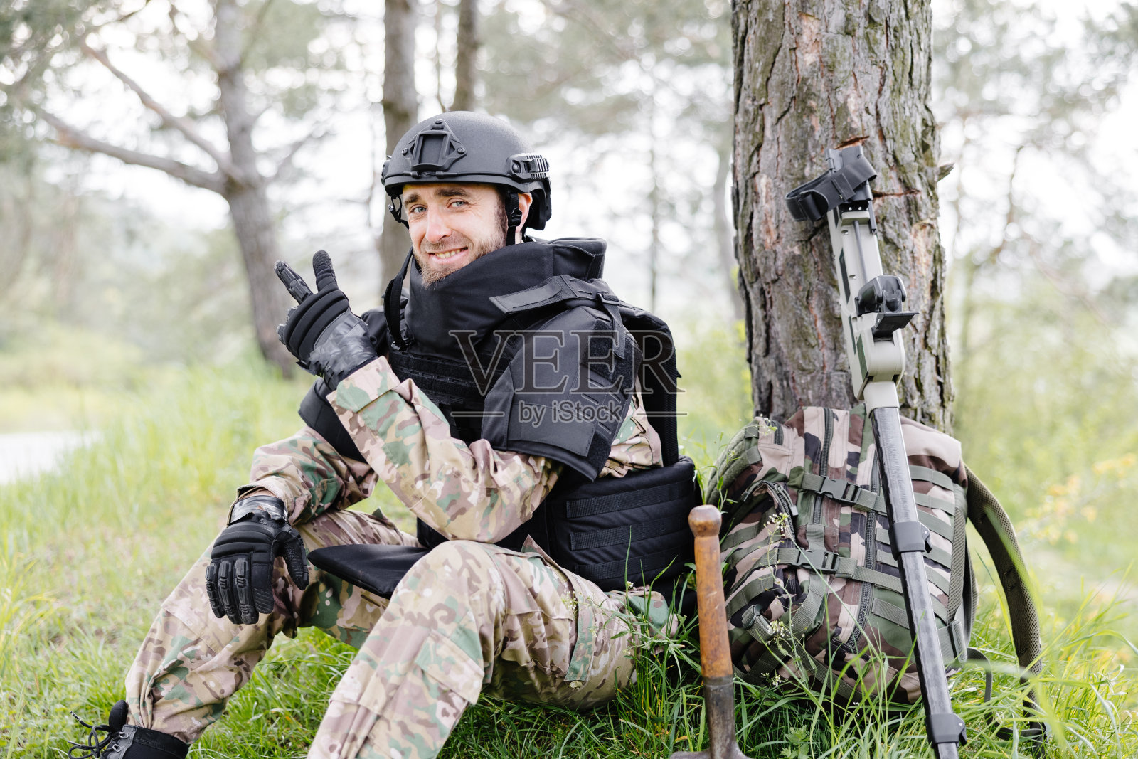 一个穿着军装和防弹背心的男人坐在森林里,旁边是一个金属探测器和一个军用背包。一名男子暂停了在该地区排雷的工作照片摄影图片