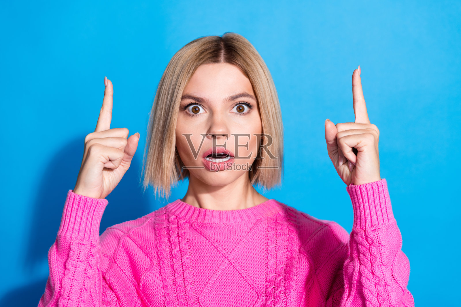照片中漂亮的年轻女子手指指向空的空间,穿着粉红色的套头衫,孤立在蓝色的背景上照片摄影图片