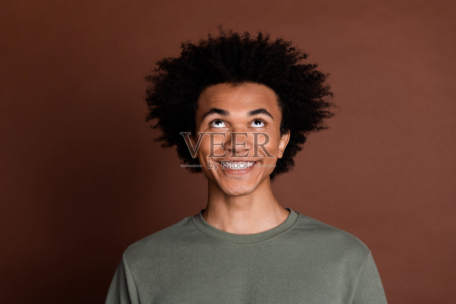 年轻时髦的家伙的肖像照片,卷发,穿着灰色t恤,微笑着抬头,在棕色背景上孤立的好主意照片摄影图片