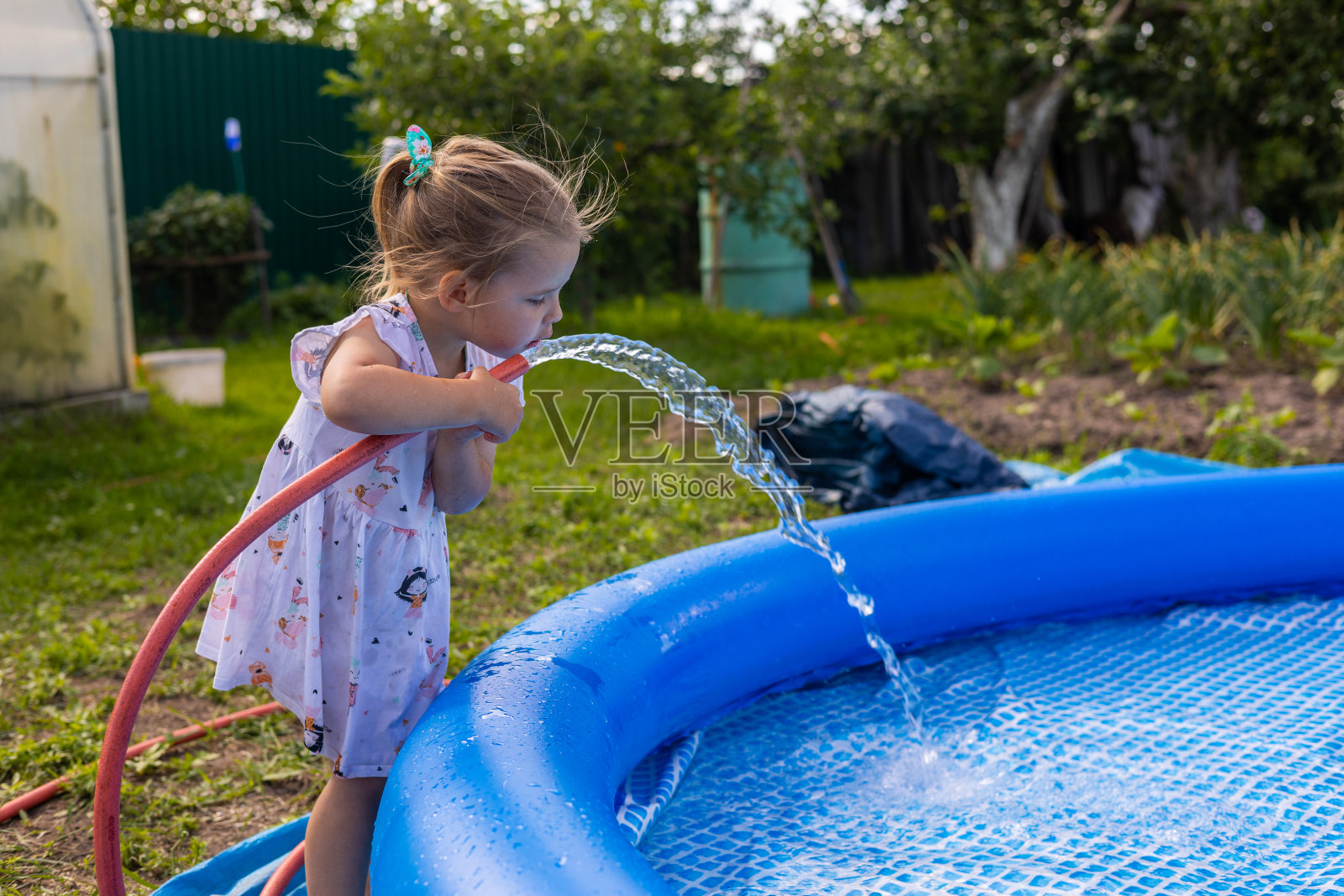 可爱的小孩用水管把水倒进池子里。一个小女孩弯下腰,从装满水池的水管里喝凉水。夏天,孩子把水池装满水。照片摄影图片