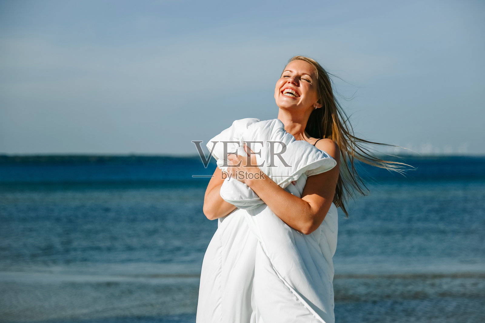 在一个阳光明媚的日子里,年轻女子站在海边,快乐地拥抱着一条毛茸茸的毯子照片摄影图片