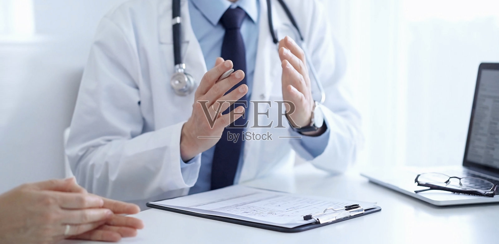 医生和病人。这名医生穿着一件白色大褂,外面是一件蓝色衬衫,打着领带,在诊所里做着手势。医学的概念照片摄影图片