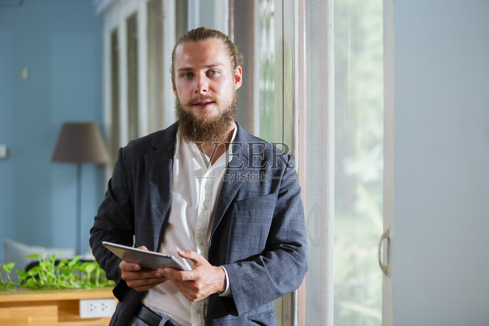 肖像英俊的商人,有胡子,拿着写字板站在办公室的侧窗。快乐聪明的男人成熟穿西装。照片摄影图片