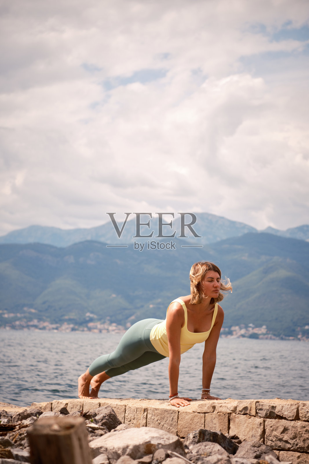 一个穿着黄色上衣和绿色打底裤的女人在岩石海岸上做了一个木板姿势,背景是平静的大海和山脉,促进核心力量和稳定性。照片摄影图片