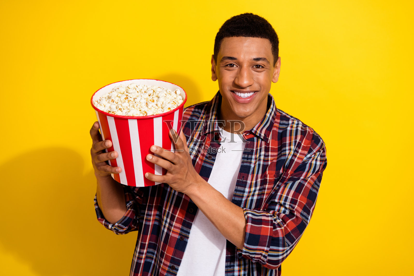 一个开朗的好男人,穿着时髦的格子衣服,看电影,在黄色的背景上看美味的爆米花照片摄影图片